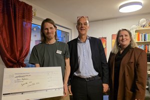 Drei Menschen lächeln in die Kamera in einem Raum. Ein Mann mit schulterlangem Haar hält einen großen Scheck in die Kamera.