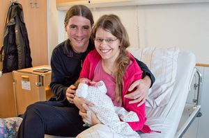 Die jungen Eltern sitzen auf dem Krankenhausbett, die Mutter hält das Baby im Arm.