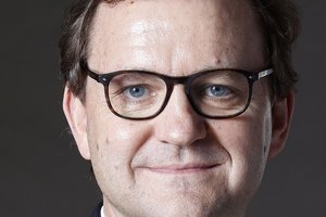 Ein Mann mit schwarzer Brille, Hemd und Jackett lächelt in die Kamera