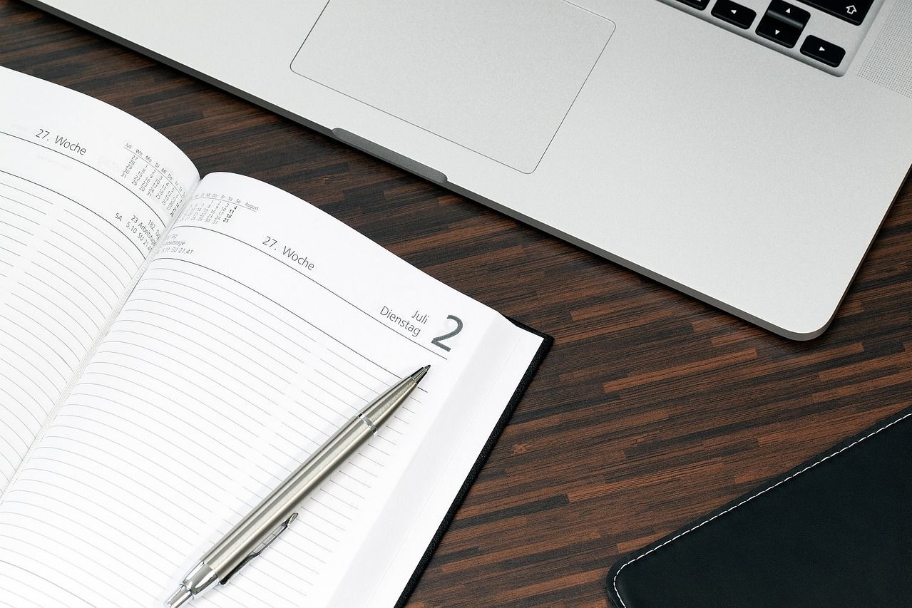Ein aufgeschlagenes Notizbuch mit einem Stift liegt auf dem Tisch, im Hintergrund ein Laptop