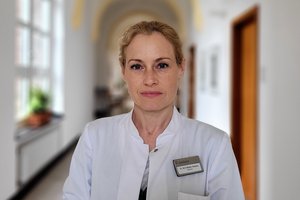 Eine blonde Frau schaut in einem weißen Arztkittel lächelnd in die Kamera auf einem Flur mit Gewölbedecke und großen Fenstern.