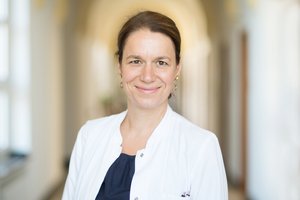 Eine Frau im Arztkittel steht auf einem warm erleuchteten Flur mit Gewölbedecke.