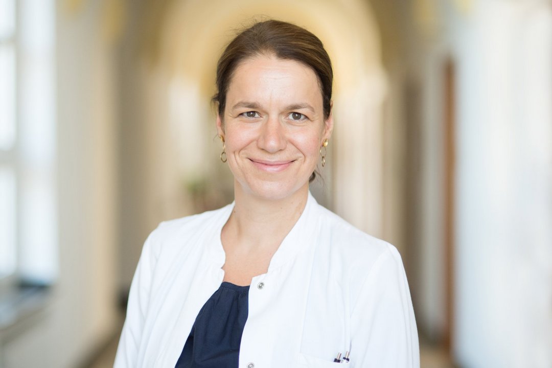 Eine Frau im Arztkittel steht auf einem warm erleuchteten Flur mit Gewölbedecke.