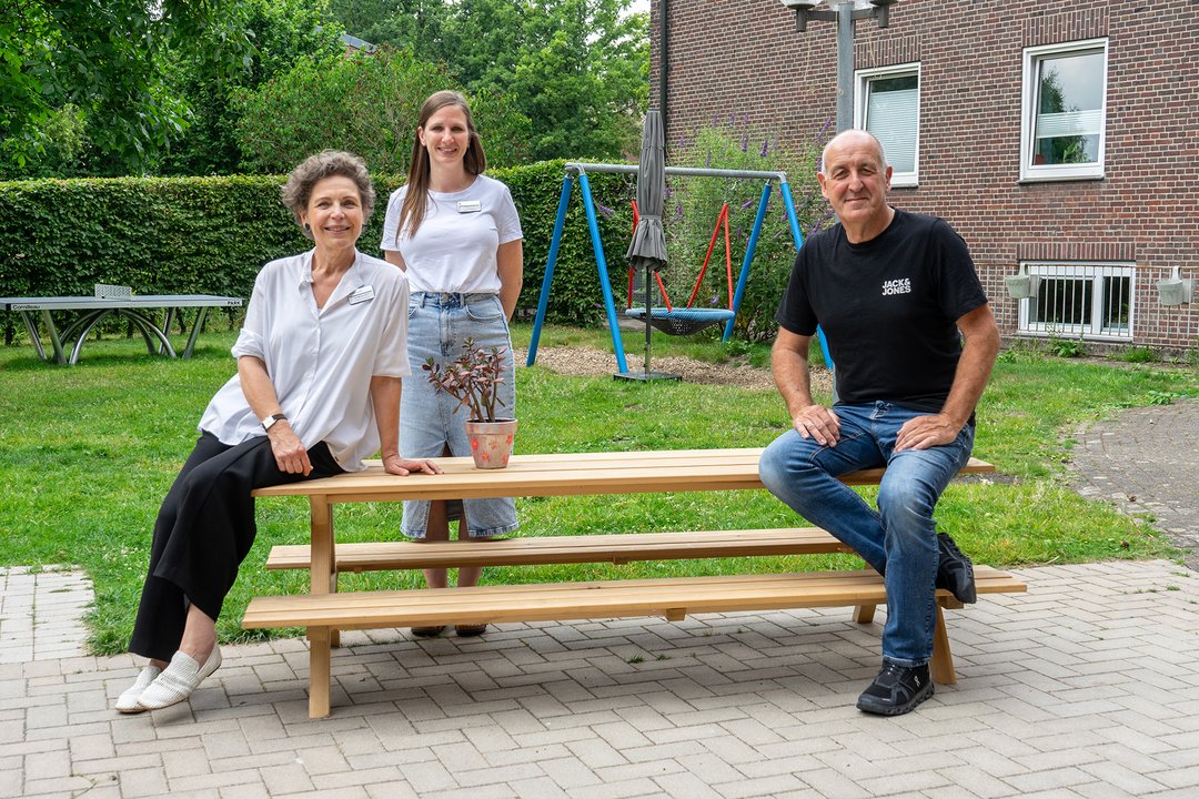 Drei Personen stehen und sitzen um einen Gartentisch herum