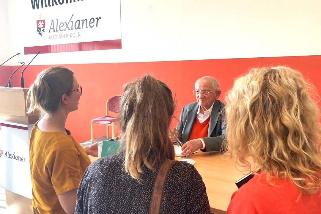 Ein älterer Mann sitzt unter dem Alexianer Logo in einer halle an einem Holztisch und wird von drei Frauen angesprochen. Er lächelt sie freundlich an. 