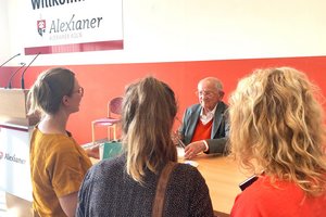 Ein älterer Mann sitzt unter dem Alexianer Logo in einer halle an einem Holztisch und wird von drei Frauen angesprochen. Er lächelt sie freundlich an. 